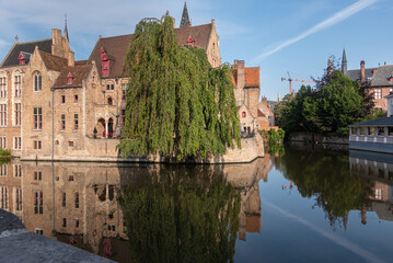 Brugge, Flanders, Belgium - August 4, 2021: Quiet Dijver reflects sunlit brown brick buildings of Wollestraat with green foliag in center and back. 