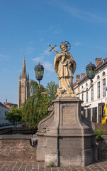 Fototapeta premium Brugge, Flanders, Belgium - August 4, 2021: Closeup of Nepomucenus stone statue on his bridge with Notre Dame cathedral tower in back under blue sky. Backdrop of green foliage and facades.