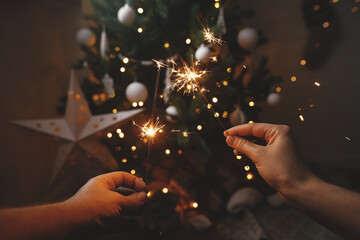 Hands holding burning sparklers on background of christmas tree and glowing star in festive scandinavian room. Happy New Year! Couple celebrating with firework bengal lights. Atmospheric moment