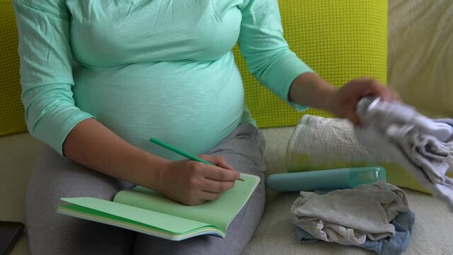 Cute Pregnant Woman writing packing list for maternity hospital With Notebook Prepares bags. Young Ledy In Pregnancy Have Fun Spend Time On Sofa At Home. Motherhood, Medicine Health and Care Concept