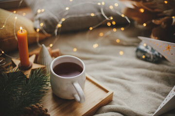 Warm cup of tea with christmas lights, stars, pine trees, candle and pillows on soft bed. Cozy home. Winter hygge. Festive holiday decorations in scandinavian evening room. Atmospheric  moment