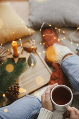 Woman hands with warm cup of tea relaxing on soft bed with golden lights, christmas stars, pine, candle and pillows in scandinavian room. Atmospheric moment. Cozy winter holidays at home. Top view