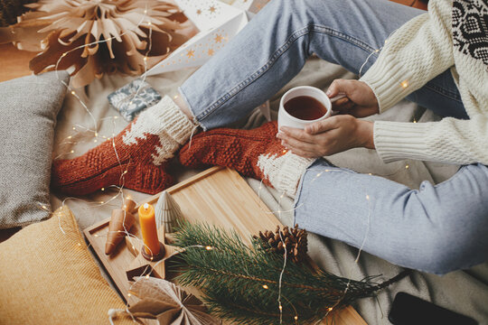 Woman hands with warm cup of tea relaxing on soft bed with festive decorations and pillows in scandinavian room with christmas lights. Cozy moments at home. Winter and autumn holidays