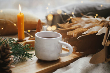 Winter hygge. Warm cup of tea with christmas lights, stars, pine trees, candle and pillows on soft bed. Cozy home. Festive holiday decorations in scandinavian evening room. Atmospheric  moment