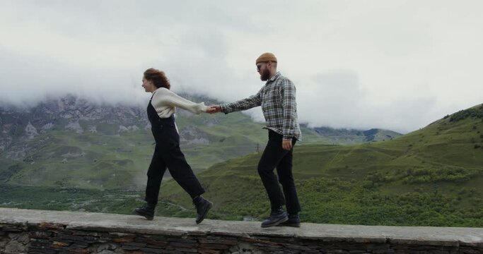 A young couple walk on the parapet holding hands against backdrop of mountains