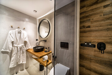 Modern tiled bathroom in beige and brown warm colors.  It has shower cabin with glass partition, wooden stand for black sink and round mirror on the wall. 