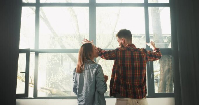 Bright future together. Rear view happy young romantic couple talk standing at big light window in new spacious home.