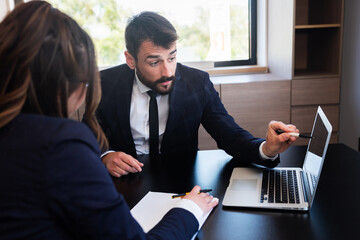 Handsome entrepreneur sitting at his desk, pointing at laptop screen and young brunette writing on paper.