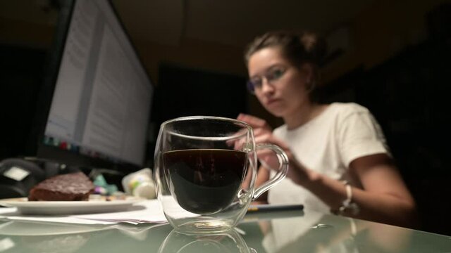 A young woman works at the computer at night and drinks coffee. Work at night and poor nutrition, not a healthy lifestyle