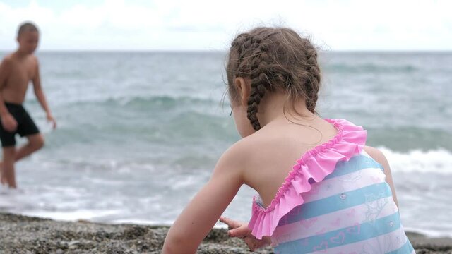 A little 3-year-old girl is sitting by the sea on a bright sunny day and playing with pebbles on a pebble beach. Relaxing on the beach. Sandy beach. Sea air. A child plays by the sea