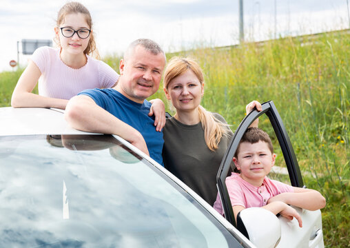 Portrait of happy parents with two teenage children posing together outdoors near car. Family travel concept.