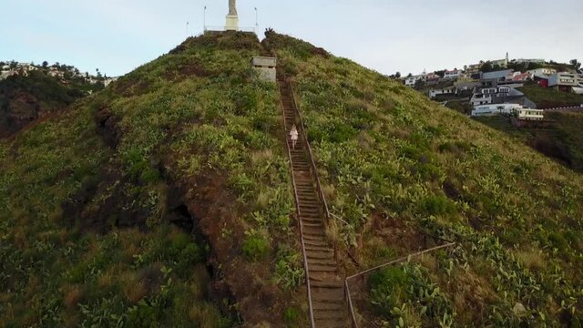 A woman walks up steep stairs to the Cristo Rei Christ Statue , Ponta do Garajau, Madeira - aerial drone shot
