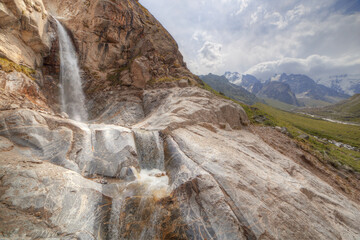 Water falls from a high cliff. In the background there are mountains with snowy peaks.
