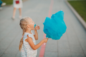 Little blond girl eating blue cotton candy in the park