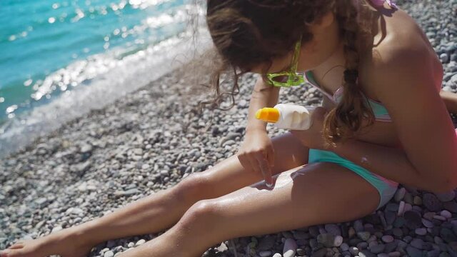 Little girl with glasses on the beach sunbathes and smears her skin with sunscreen. Kid draws on his feet with sun lotion. Travel concept, rest, vacation, tourism
