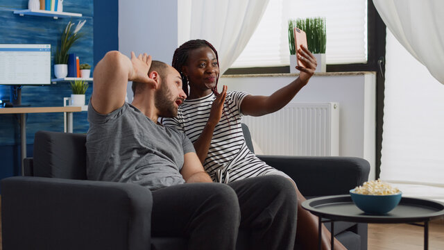 Joyful interracial couple taking pictures on smartphone while sitting in living room. Mixed race husband and wife feeling cheerful taking fun selfies and enjoying happy relationship