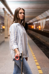 Young girl with longboard wait for train at empty subway metro station. Casual female student look...