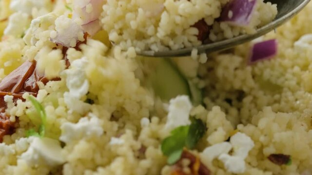 Picking Couscous salad with sun dried tomatoes, cucumber, red onion and feta cheese using a tablespoon. healthy food.