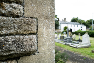 Shallow focus of an ancient stone wall seen near the entrance to an English church. The background shows some old gravestones and beyond a large house at the centre of a rural village.