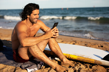 Young man using the phone while sitting on the beach. Handsome man resting after surfing