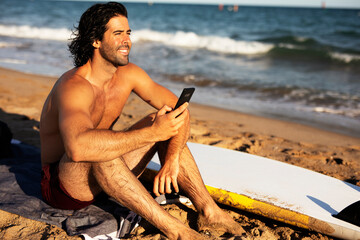 Young man using the phone while sitting on the beach. Handsome man resting after surfing