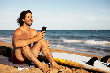 Young man using the phone while sitting on the beach. Handsome man resting after surfing