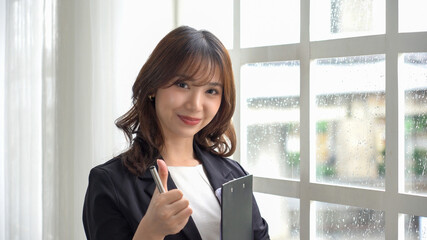 An Asian woman thumbs up and holding a note near a window on a rainy day (Chinese, Japanese, Korean or Thai) signifying success.