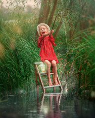 A little girl in a straw hat and red dress stands sitting on a ladder in the river with a fishing...