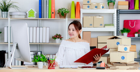 Asian young happy successful online selling businesswoman checking customer address from computer monitor and folder in hand at working table full of cardboard package shipping boxes at home office