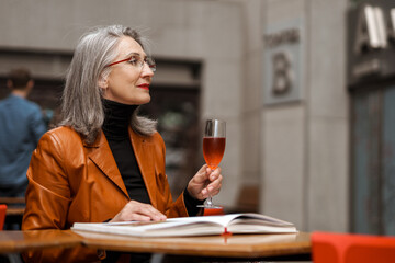 Grey white senior woman reading book while drinking wine in cafe