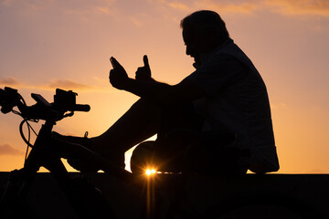 Fototapeta premium Silhouette of mature man sitting close to his bicycle at sunset light using mobile phone. Horizon over water
