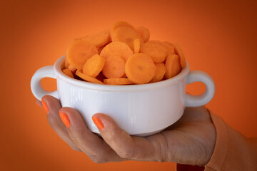 Close up on a woman's hand holding a white bowl full of round sliced carrots. Orange background