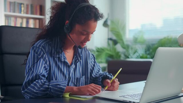 Young Indian woman with headset sitting at home getting education online. Teenage girl learns improves her knowledge of foreign language and acquires knowledge on Internet remotely using laptop.