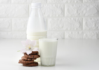 a full glass cup of milk and a clear plastic bottle with milk, next to a stack of round chocolate chip cookies on a white table