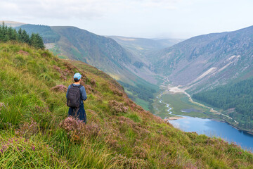 Naklejka na ściany i meble Young man admiring the view over the Glendalough Lake and Glenealo Valley, in Wicklow Mountains, Ireland, after hiking a segment of the popular Spinc trail.