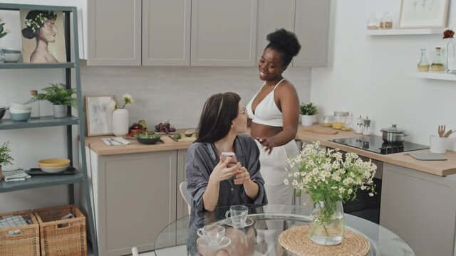 Handheld slowmo shot of African-American woman smiling and pouring coffee for her Caucasian girlfriend sitting at table in cozy kitchen