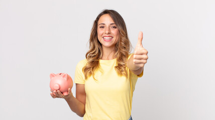 pretty thin woman feeling proud,smiling positively with thumbs up and holding a piggybank