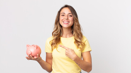 pretty thin woman smiling cheerfully, feeling happy and pointing to the side and holding a piggybank - obrazy, fototapety, plakaty