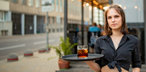 Portrait young waitress standing in outdoor cafe. girl the waiter holds a tray with a glass of...