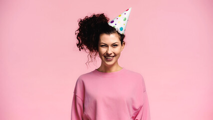 cheerful curly woman in party cap smiling at camera isolated on pink