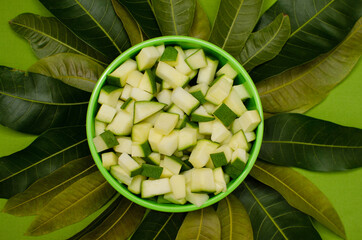 Raw mango pieces in Green bowl with mango tree leaves on Green background