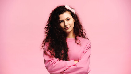 Fototapeta premium positive woman standing with crossed arms and smiling at camera isolated on pink