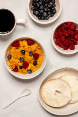 Continental breakfast of cereal, bagel, cornflakes, raspberry, blueberry and coffee on white marble in a vertical orientation