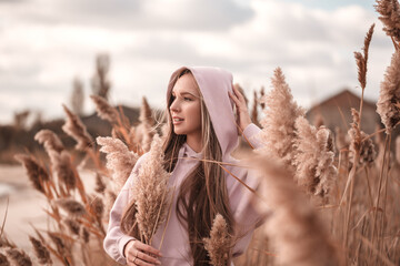 Autumn portrait of a young beautiful woman in a warm knitted hoodie outside among the dry reeds. Cozy photo toned in warm colors.