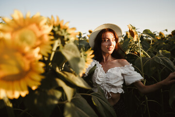 Smiling woman wearing a hat standing in a field of sunflowers in sunset