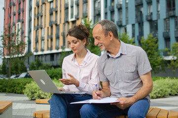 Attractive young woman and senior man are using laptop sitting on the bench in modern residential complex. Casual meeting outside office. A daughter is teaching technology for her father