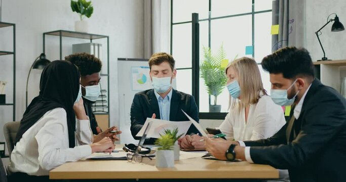 Handsome experienced ceo in protective mask holding business meeting with his confident different ages mixed race subordinates in protective masks too,office work during pandemia of covid-19