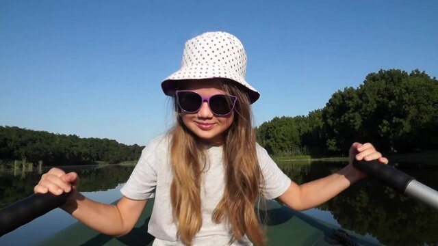 Little smiling child girl in sunglasses and hat rowing along the river. Child controls the boat independently. Kid sails on a boat outdoors on summer vacation. Fishing with dad