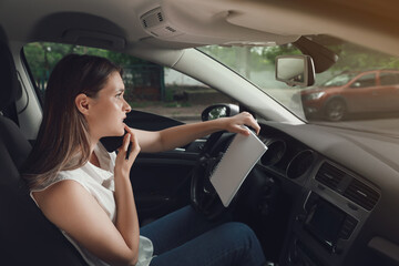 Fototapeta na wymiar Stressed young woman with notebook talking on phone in driver's seat of modern car