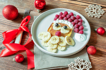 Christmas Santa Claus shaped pancake with sweet fresh raspberry berry and banana on plate on wooden background for kids children breakfast. xmas food with new year decorations close up